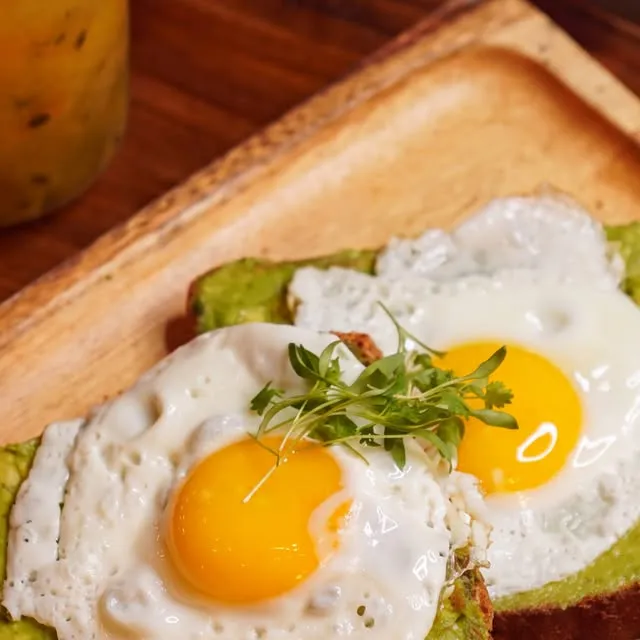 Dos rebanadas de pan con aguacate y huevos estrellados sobre una tabla de madera, con fruta.