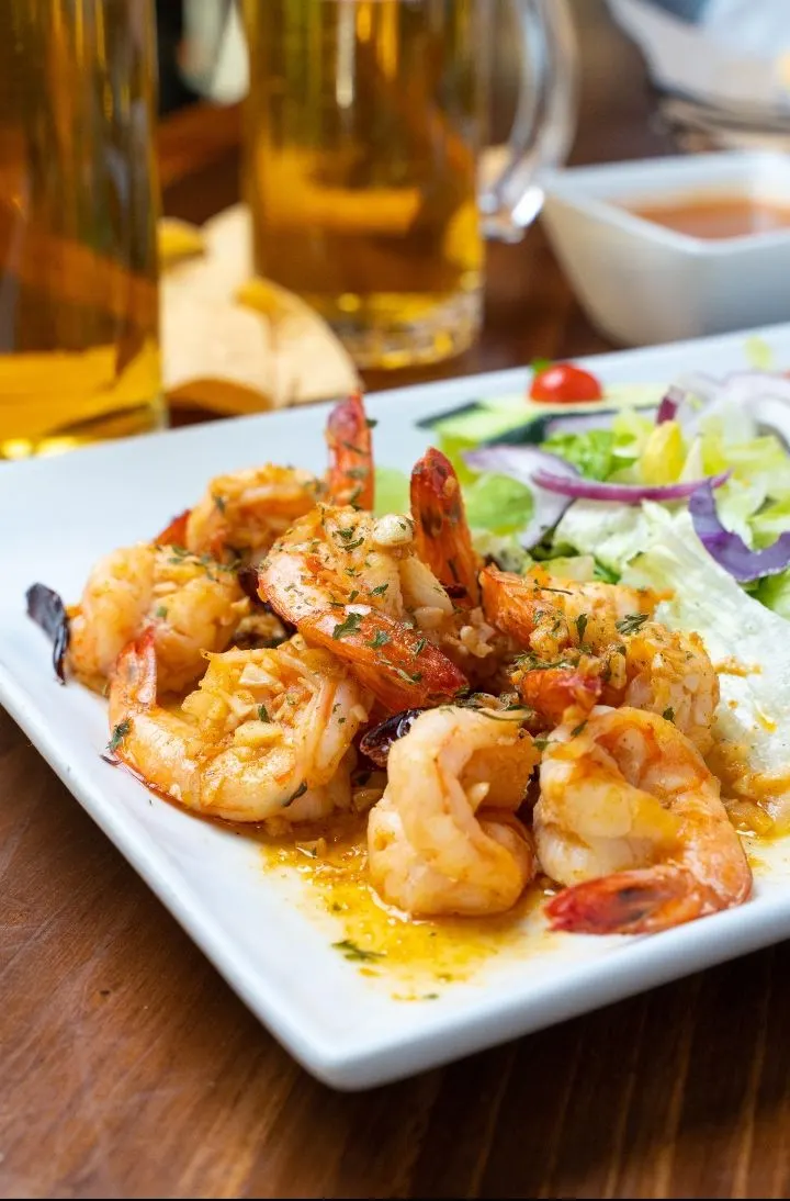 Camarones al ajo en un plato blanco con ensalada y un tarro de cerveza al fondo.