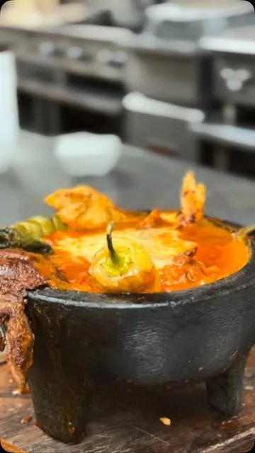 Un molcajete de piedra con salsa roja burbujeando, camarones, carne asada y un chile güero.