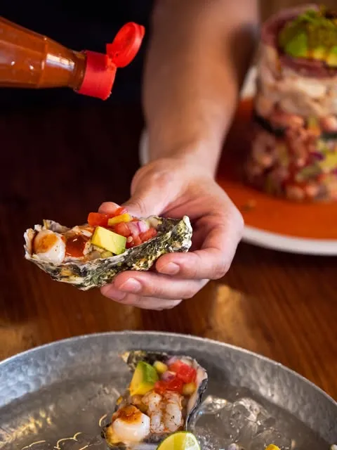 Una mano sostiene un ostión preparado con camarón y aguacate; hay más en la charola.